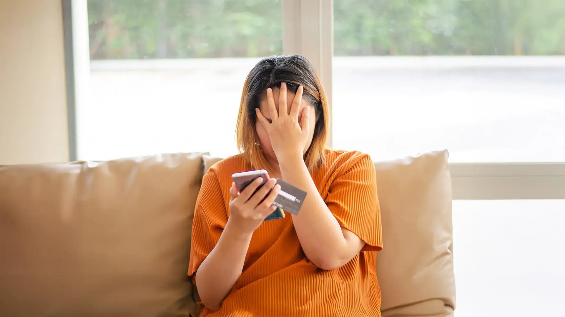 Mujer sentada cubriéndose el rostro con preocupación mientras sostiene un teléfono móvil y una tarjeta bancaria.