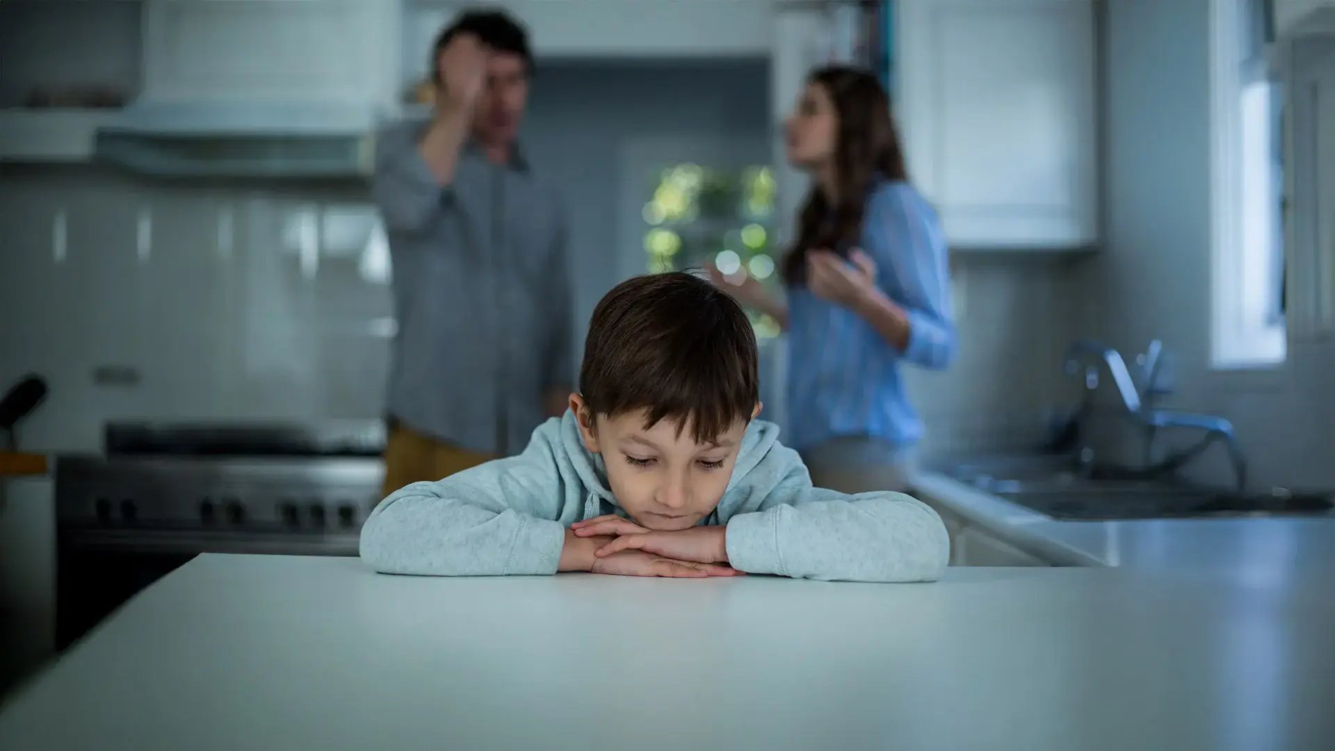 Niño pequeño sentado solo y triste en una mesa, con figuras de adultos desenfocadas discutiendo al fondo.